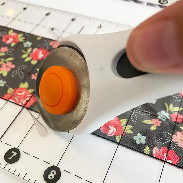 Rotary fabric cutter with an orange safety button, being used to cut floral-patterned fabric.